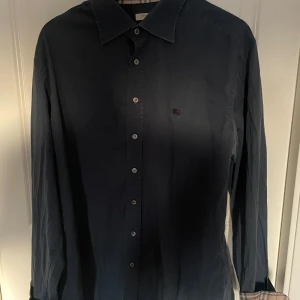 Mörkblå skjorta från Burberry XL - mörkblå skjorta från Burberry London i storlek XL. Klassisk modell med knappar framtill, lång ärm och ikoniskt rutigt tyg på insidan av kragen och vid manchestterna. Finns i ett djur och rökfritt hem