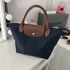 Blå handväska från Longchamp - Snygg marinblå handväska från Longchamp med bruna skinnhandtag och lock. Väskan har klassisk form, guldknapp och är tillverkad i slitstark syntet med detaljer i skinn. Perfekt storlek för vardagens essentials och enkel att matcha till olika outfits.