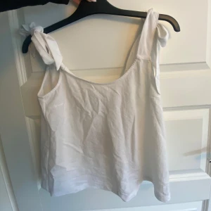 Vit linnetopp med knytaxlar - Superfin vit linnetopp med breda axelband som knyts i stora rosetter på axlarna. Toppen har en avslappnad passform och är tillverkad i ett svalt material, perfekt för varma dagar. Enkel och stilren design som är lätt att matcha med allt.
