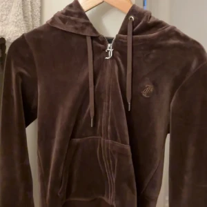 Brun velour hoodie från Juicy Couture - Mjuk och lyxig brun hoodie från Juicy Couture i velour med dragkedja framtill, huva och snörning. Klassisk logga broderad på bröstet och känguruficka. Perfekt för en chill och trendig look. 