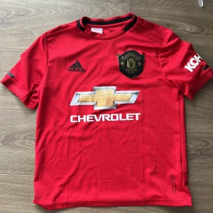 Manchester United Adidas matchtröja  - Säljer en röd Manchester United fotbollströja från Adidas. Tröjan har korta ärmar, svart klubbmärke och sponsorloggor på bröstet och ärmen. Tillverkad i lätt och ventilerande polyester med Climalite-teknologi. Perfekt för dig som älskar fotboll och United! Storlek 164
