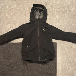 Svart vindjacka från Haglöfs med huva - Svart vindjacka från Haglöfs med huva, hel dragkedja och stora fickor framtill. Jackan har justerbar nederkant med snörning och reflexdetaljer på ärmarna. Perfekt för blåsiga dagar och outdoor-äventyr. Snygg och funktionell design. Storlek 123