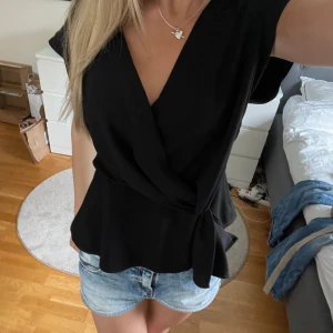 Zara Omlottopp - Snygg svart omlottopp med v-ringning och knyt i midjan. Toppen har korta ärmar och en lätt utsvängd nederdel som ger en fin siluett. Perfekt att styla med jeansshorts eller kjol för en trendig look.