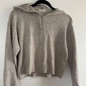 Beige stickad hoodie från JDY - Mysig beige stickad hoodie från JDY med huva och snörning. Tröjan har ribbade muddar och är lite croppad i modellen. Perfekt för lager-på-lager och chill dagar. Materialet är en mjuk blandning med polyester.
