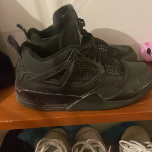 Svarta Nike Air Jordan 4 sneakers - Säljer ett par helsvarta Nike Air Jordan 4 sneakers med klassisk meshpanel på sidorna, snörning och ikonisk Jumpman-logga på plösen. Skorna har en chunky sula och coola detaljer i syntet och skinn för en riktigt streetig vibe. de är i helt okej skick. Jag har använt dem i några månader.