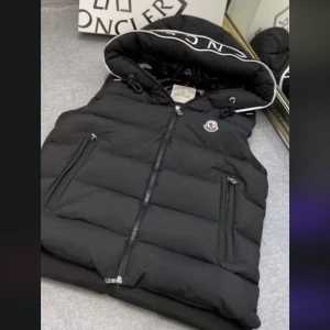 Svart väst Moncler - Snygg svart dunväst från Moncler med stor huva och vit logotyptext längs kanten. Västen har två dragkedjefickor, quiltad design och klassisk Moncler-logga på bröstet. Perfekt för lager-på-lager och streetstyle. Stängs med dragkedja framtill. Vid frågor skriv privat må bäst 