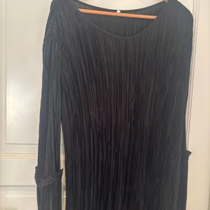 Svart plisserad långklänning - Snygg svart långklänning med plisserat tyg och långa ärmar. Klänningen har en rund halsringning och coola detaljer med veckade partier vid ärmsluten. Perfekt för dig som gillar stilrena och moderna vibes.