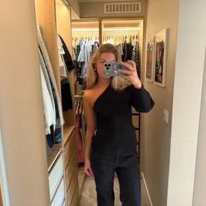Svart oneshoulder topp från Zara - Säljer en svart, stilren oneshoulder topp från Zara. Toppen har en lång ärm på ena sidan och är ärmlös på andra. Materialet är stretchigt och sitter snyggt på kroppen. 