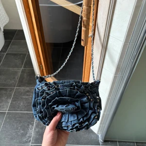 Jeans axelväska med volang och nitar - Säljer en unik axelväska i jeansmaterial med volangdetaljer framtill och små silvriga nitar. Väskan har en kedjerem som axelband och stängs med magnetknapp. Perfekt för dig som vill sticka ut med en cool och trendig accessoar.