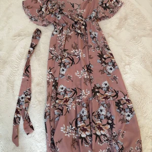 Blommig långklänning från SHEIN -  långklänning från SHEIN i en mjuk rosa ton med blommigt mönster i vitt, beige och svart. Klänningen har v-ringning, volangärmar och avtagbart midjebälte.  flowy passform och lätt material som känns luftigt.