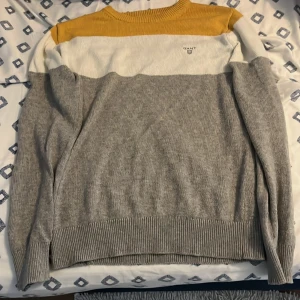 GANT färgblockad stickad tröja - Säljer en snygg stickad tröja från GANT med färgblock i gult, vitt och grått. Tröjan har rund halsringning, långa ärmar och diskret GANT-logga på bröstet. Perfekt för dig som gillar stilren men ändå färgglad look. Storleken är Xs men sitter också ganska bra på mig som har storlek S