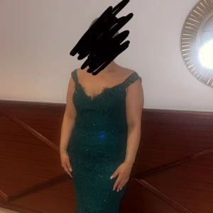 Grön lång spetsklänning med paljetter - Säljer en elegant lång grön klänning med v-ringning och offshoulder. Klänningen är täckt av spets och glittriga paljetter som ger en lyxig känsla. Perfekt för bal eller fest. Sitter figurnära och har en snygg passform.