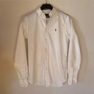 Vit skjorta från Ralph Lauren slim fit - Klassisk vit skjorta från Ralph Lauren i slim fit-modell. Skjortan är i lätt bomullstyg, har långa ärmar, knappar framtill och button down-krage. Broderad logga med polospelare på bröstet ger en clean och tidlös look.