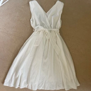 Vit klänning med knytband - Superfin vit omlottklänning med v-ringning och knytband i midjan. Klänningen är ärmlös och har en luftig, flowy passform som passar perfekt till sommaren..