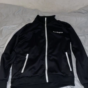 Svart kofta Black Squad S - Svart kofta från Black Squad med vita detaljer och dragkedjor. Kofta har ståkrage, vita ränder längs ärmarna och texten 'Los Angeles' broderad på bröstet. Perfekt för en sportig och chill stil.