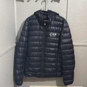Svart pufferjacka EA7 Emporio Armani - Snygg svart pufferjacka från EA7 Emporio Armani med huva och dragkedja framtill. Jackan har ett stilrent, quiltat mönster och logga på bröstet. Tillverkad i lätt syntetmaterial och levereras med tillhörande förvaringspåse. Perfekt för kalla dagar.
