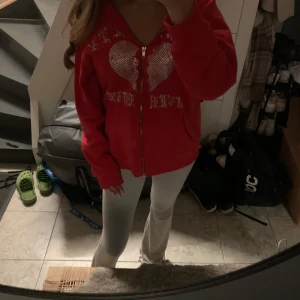 Röd hoodie med strasshjärta och dragkedja - Röd hoodie med dragkedja framtill och stor strassdekor i form av ett brustet hjärta på bröstet. Text i strass ovanför och under hjärtat. Skriv vid frågor❤️( passar s)