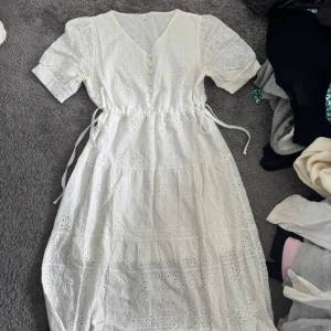 Vit långklänning med spets och pärlknappar - Superfin vit långklänning med broderade spetsdetaljer och pärlknappar framtill. Klänningen har v-ringning, korta ärmar och knytband i midjan för en snygg passform. Perfekt för sommarens alla tillfällen!