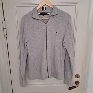 Grå kofta från Tommy Hilfiger M - Säljer en stilren grå kofta från Tommy Hilfiger i storlek M. Koftan har hel dragkedja framtill, ribbade muddar och diskret logga på bröstet. Tillverkad i mjuk bomull och passar perfekt till lager-på-lager. Klassisk krage med Hilfigers signaturränder.
