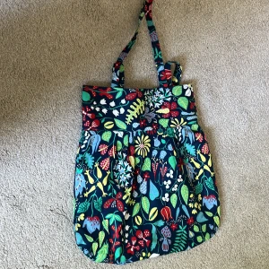 Färgglad blommig shoppingväska - En stor och rymlig shoppingväska i bomull med färgglatt blommönster i rött, gult, grönt, blått och vitt mot mörkblå botten. Väskan har dubbla axelband och är perfekt för att bära med sig allt man behöver. Snygg och unik accessoar för dig som gillar färg och mönster.