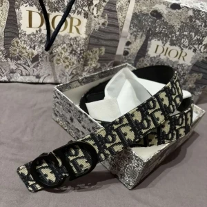 Svart och beige Dior bälte med logomönster - Knappt använd bälte från Dior med ikoniskt svart och beige logomönster över hela bandet. Bältet har en stilren svart spänne med CD-detalj och är tillverkat i canvas med skinn på insidan. Perfekt accessoar för att lyfta din outfit med lyxig känsla och tydlig designerstil.