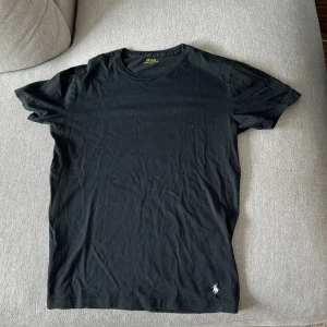 Svart t-shirt från Polo Ralph Lauren - Svart t-shirt från Polo Ralph Lauren i klassisk modell med kort ärm och rund hals. Tillverkad i mjuk bomull och prydd med den ikoniska broderade loggan i vitt vid nederkanten. Enkel och stilren, perfekt till jeans eller shorts.