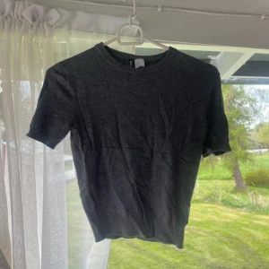 Mörkgrå basic kortärmad stickad topp från H&M - En stilren mörkgrå kortärmad stickad topp  i klassisk modell. T-shirten har rund halsringning, korta ärmar och är tillverkad i mjuk bomull. Perfekt till jeans eller shorts för en enkel och clean look.