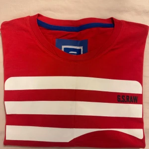 Röd med vit randig t-shirt från G-Star RAW - Snygg röd t-shirt från G-Star RAW med vita horisontella ränder och tryckt logga på bröstet. Klassisk rund halsringning och korta ärmar. Materialet är mjuk bomull och det finns en broderad G-Star Originals patch på ärmen. Perfekt för en cool och avslappnad stil.
