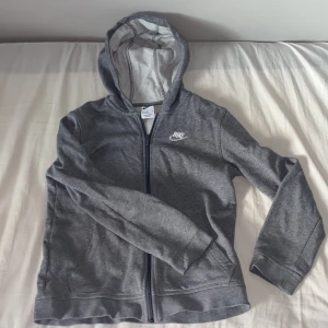 Grå Nike hoodie med dragkedja - Snygg grå hoodie från Nike med broderad logga på bröstet. Tröjan har huva, hel dragkedja och två fickor framtill. Perfekt för chill dagar eller när du vill ha en sportig look. Mjuk bomullsblandning som känns skön mot huden.
