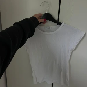 Vit ribbad topp från Zara - Enkel och stilren vit ribbad topp från Zara. Modellen är ärmlös med rund halsringning och har en tight passform. Perfekt att matcha med jeans eller kjol för en clean look. Materialet är mjukt och stretchigt för extra komfort.