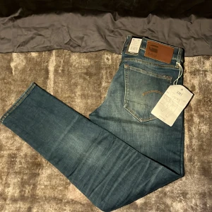 G-Star RAW 3301 blå jeans - Snygga blå jeans från G-Star RAW, modell 3301. Klassisk femficksdesign med läderpatch bak och tydliga slitningar framtill för en cool look. Jeansen har normal passform och raka ben. Tillverkade i slitstarkt denim med orange kontrastsömmar.