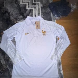 Vit långärmad Frankrike Nike träningströja - Vit långärmad träningströja från Nike för Frankrikes landslag. Guldiga detaljer med broderad tupp och två stjärnor på bröstet, samt Nike-logga. Halvlång dragkedja och diskreta mönster på ärmarna. Tillverkad i lätt och ventilerande funktionsmaterial.