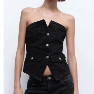 Svart jeans tubtopp med knappar - Säljer en svart axelbandslös tubtopp i jeansmaterial med coola silverfärgade knappar framtill och två fickdetaljer. Toppen har en unik v-ringad skärning upptill och är croppad för en edgy look.