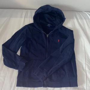  Polo Ralph Lauren zip hoodie - Snygg marinblå hoodie från Polo Ralph Lauren med dragkedja och klassisk röd broderad logga på bröstet. Tröjan har huva, känguruficka och är tillverkad i mjuk bomull. Perfekt för en chill och stilren look.