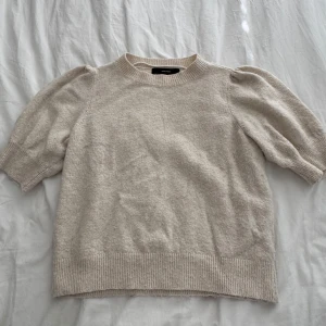 Beige stickad tröja med puffärm - Superfin beige stickad tröja från Vero Moda med korta puffärmar och ribbade detaljer vid hals, ärmslut och nederkant. Perfekt för lager-på-lager och ger en chill men ändå söt vibe till din outfit.