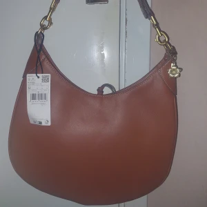 Brun axelväska från Mango i skinnimitation - Helt ny - Säljer en snygg brun axelväska från Mango med guldfärgade metalldetaljer. Väskan har en mjukt rundad form och är gjord i skinnimitation, den har ingen dragkedja utan man knyter ihop banden för att stänga. Perfekt storlek för det viktigaste och enkel att matcha till olika outfits.