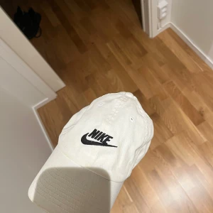 Vit keps från Nike med svart logga - Snygg vit keps från Nike med klassisk svart broderad logga framtill. Kepsen har böjd skärm och justerbar passform bak. Tillverkad i bomull för en skön och luftig känsla. Perfekt accessoar för en sportig och avslappnad stil.