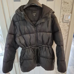 Svart jacka från Lindex - Säljer en svart jacka från Lindex med huva och justerbar midja. Jackan har dragkedja framtill.
