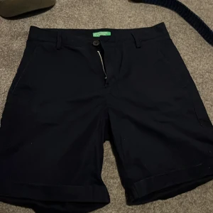 Svarta shorts från United Colors of Benetton - Klassiska svarta shorts från United Colors of Benetton med snyggt uppvikta benslut, fickor fram och bak samt bälteshällor. Perfekta för sommaren och har en stilren look som funkar till många outfits.