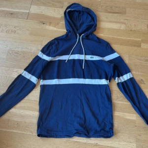 Blå randig hoodie från Lacoste - Snygg blå hoodie från Lacoste med vita horisontella ränder och klassisk krokodillogga på bröstet. Tröjan har huva med snörning och långa ärmar. Tillverkad i mjuk bomull, perfekt för en chill och sportig stil.