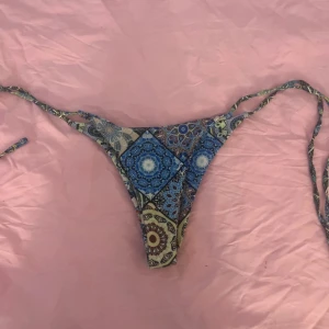 Blå mönstrad bikiniunderdel med snörning - Säljer en snygg bikiniunderdel med blått och beige mönster i boho-stil. Modellen har högt skurna ben och smala knytband i sidorna för en justerbar passform. Perfekt för strandhäng och soliga dagar.