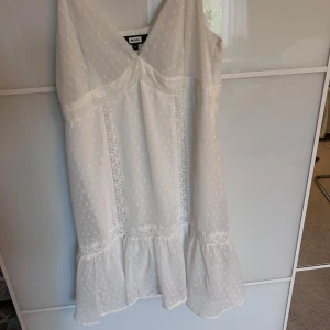 Vit kortklänning med spets från Bik Bok - Supersöt vit kortklänning från Bik Bok i storlek XS. Klänningen har tunna axelband, v-ringning och är dekorerad med spetsdetaljer och diskret prickigt mönster. 