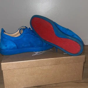 Blå mocka sneakers från Louboutin - Snygga blå sneakers i mocka från Christian Louboutin med ikonisk röd sula. Skorna har rund tå, platt sula och klassisk snörning. Insidan är ljusbeige och designen är stilren men ändå riktigt iögonfallande tack vare färgkombon. Kan handla om priset 