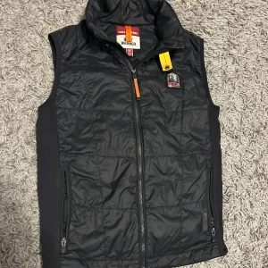 Svart dunväst från Parajumpers - Svart dunväst från Parajumpers med hög krage, dragkedja framtill och två sidofickor med dragkedja. Västen har en gul detalj vid halsen och Parajumpers-logga på bröstet. Perfekt för lager-på-lager och sommarkvällar. Tveka inte på att skriva vid minsta fundering.