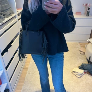 Svart majedupe - Säljer en svart fyrkantig handväska från Don Donna  med coola fransar på sidorna. Väskan har både handtag och avtagbar axelrem så du kan bära den på flera sätt. Perfekt storlek för mobilen, plånboken och lite till.