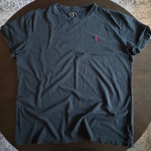 Svart t-shirt från Ralph Lauren - En klassisk svart Ralph Lauren t-shirt med rött tryck på bröstet. Bra skicka och passar till nästan allt, speciellt nu till sommaren. Om du har några funderingar så är det bara att fråga på.