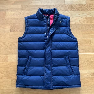 Blå dunväst från Gant - Snygg blå dunväst från Gant med quiltad design och dragkedja. Perfekt för kyliga dagar med sin stilrena look och bekväma passform. Västen har praktiska fickor och en diskret logga på bröstet. Nyskick! 