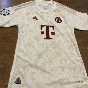 Beige fotbollströja från Adidas - Säljer en snygg beige fotbollströja från Adidas med ett subtilt blommönster. Tröjan har korta ärmar och ett tryck i vinrött på bröstet. Perfekt för fotbollsfans som vill ha något unikt! 🌟