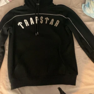 Svart hoodie från Trapstar - Säljer en svart hoodie från Trapstar med vit text på bröstet. Hoodien har långa ärmar och en justerbar huva med dragsko. Tillverkad i en bomulls- och polyesterblandning för komfort och hållbarhet.