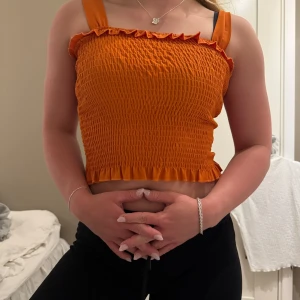 Orange ribbad linnetopp - Säljer en snygg orange ribbad linnetopp med volangdetaljer vid axelband och nederkant. Perfekt för sommardagar och ger en fräsch look. Toppen är croppad och har en tight passform.